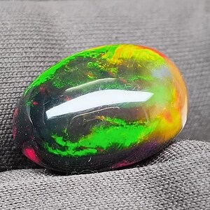 May include: A polished, oval-shaped opal gemstone. The opal displays a vibrant play of color, with flashes of green, yellow, red, and black. The gemstone is set against a dark gray fabric background, highlighting its iridescent qualities.