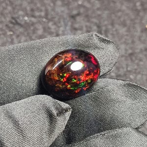 Könnte beinhalten: Ein dunkler ovaler Opal-Edelstein mit roten, grünen und schwarzen irisierenden Blitzen.