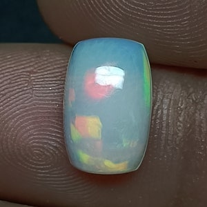 May include: A white opal cabochon with a light blue, green, and pink iridescent sheen. The opal is oval shaped and has a smooth, polished surface.