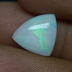 Könnte beinhalten: Ein dreieckiger, weißer Opal-Edelstein mit schillernden grünen und blauen Blitzen. Der Opal ist im Cabochon-Schliff gefasst.