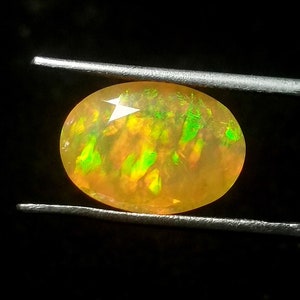 Könnte beinhalten: Ein gelber Opal-Edelstein mit grünen und orangefarbenen Farbschimmern. Der Opal ist oval geformt und wird von einer Pinzette gehalten.