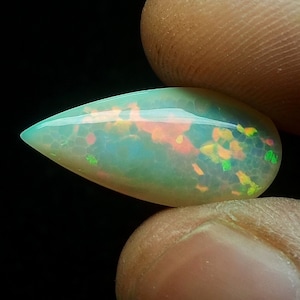 May include: A teardrop-shaped opal gemstone with a translucent, iridescent appearance. The opal displays a mix of colors, including green, orange, and yellow, with a network of internal patterns. The gemstone is held between two fingers against a black background.