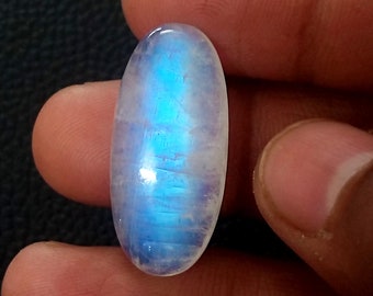Könnte beinhalten: Ovaler Mondstein-Cabochon mit einem blauen, irisierenden Schimmer. Der Stein ist durchscheinend und hat eine milchig weiße Farbe.
