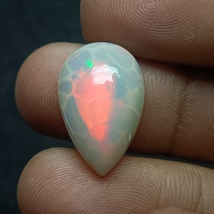 Könnte beinhalten: Ein tropfenförmiger Opal-Edelstein mit einer weißen Basis und einem rot-grünen, schillernden Schimmer.