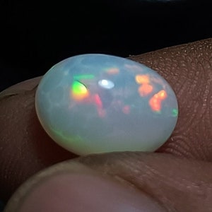 May include: A polished, oval-shaped opal gemstone. The opal has a milky white base color with flashes of orange, red, and green iridescence. The gemstone is held between fingers against a dark background.