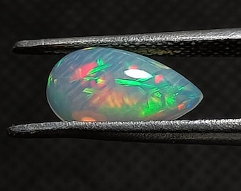 Könnte beinhalten: Ein perlmuttartiger weißer Opal-Edelstein mit grünen, roten und orangefarbenen Farbschimmern. Der Opal hat die Form eines Tropfens und wird von einer Pinzette gehalten.