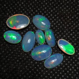 May include: A collection of eight oval-shaped opals with iridescent colors, including green, blue, and orange. The opals are arranged on a dark gray textured surface.