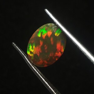 Könnte beinhalten: Nahaufnahme eines grünen, roten und orangefarbenen Opals, der in einer Pinzette gehalten wird. Der Opal ist oval geformt und hat eine glatte, polierte Oberfläche.