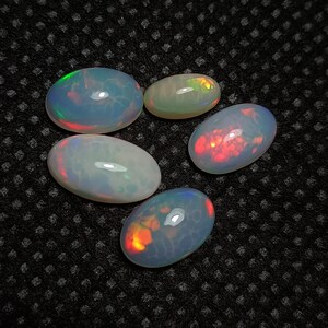 May include: Five oval-shaped opals with iridescent colors, including red, orange, green, and blue. The opals are arranged in a semi-circle on a black textured surface.