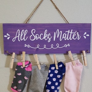 May include: A purple wooden sign with the text "All Socks Matter" in white script. The sign has a jute rope hanger and wooden clothespins holding up four pairs of socks in various colors and patterns.