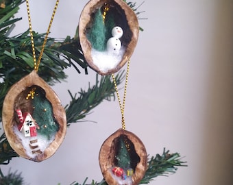 Può includere: Tre decorazioni natalizie fatte a mano appese a un albero verde. Ogni decorazione è un guscio di noce con una scena invernale in miniatura all'interno, tra cui un pupazzo di neve, una casa e un albero di Natale. Le decorazioni sono sospese da cordoncini dorati.