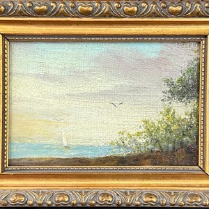 Puede incluir: Una pintura al óleo sobre lienzo que representa un paisaje costero con un velero en aguas azules, una orilla con árboles verdes y un cielo pálido con pájaros, encerrada en un marco rectangular dorado y ornamentado con detalles en relieve.