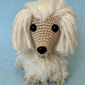 May include: A handmade crochet dog with a cream-colored body and head, featuring black eyes and a black nose. The dog has fluffy, cream-colored yarn around its head and body, giving it a soft appearance. The background is a solid light blue.