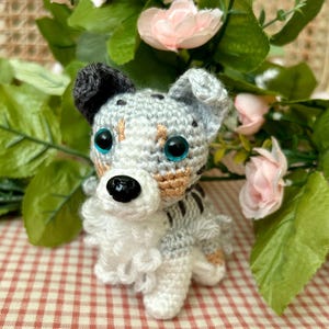 May include: A crocheted stuffed dog toy with blue eyes, a white chest, and brown and gray spots. The dog is sitting on a red and white checkered surface.