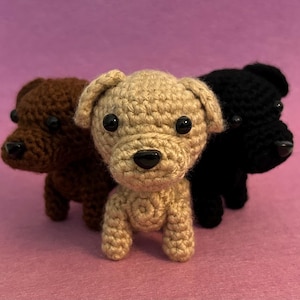 May include: Three crocheted dog figurines in brown, tan, and black. Each dog has black button eyes and a black nose. The dogs are standing on a pink surface.