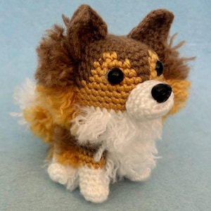 Pode incluir: Uma figura de cão de crochê em tons de marrom, bege e branco. O bicho de pelúcia feito à mão apresenta olhos de botão pretos e um nariz preto. O cão tem uma juba e cauda fofas e está posicionado em uma superfície azul clara.