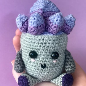 Puede incluir: Un juguete de peluche tejido a crochet con forma de planta suculenta en una maceta. La planta es morada y gris, y la maceta es gris. El juguete tiene una cara linda con ojos negros y un rubor rosado.