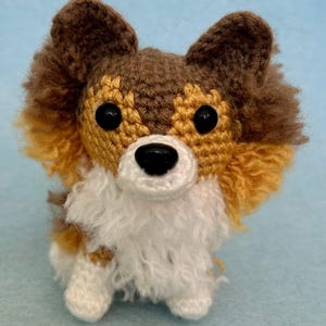 May include: A handmade crochet dog with brown, tan, and white yarn. The dog has black button eyes, a black nose, and fluffy fur around its face and chest. The dog is sitting on a light blue surface.
