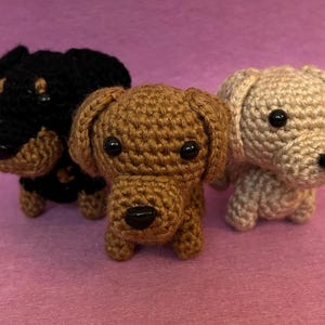 May include: Three crocheted dog figurines in black, brown, and tan. Each dog has black button eyes and a black nose. The dogs are standing on a pink surface. The dogs are handmade.