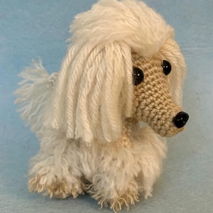 Peut inclure: Un caniche au crochet fait à la main, assis. Le chien a un corps et une tête duveteux de couleur crème, avec un museau crocheté beige. Des yeux en boutons noirs et un nez noir complètent le design. La fourrure du caniche est en fil doux.