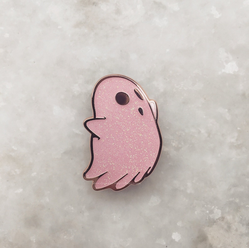 May include: A pink glitter ghost enamel pin. The ghost has a simple, cartoon-like design with a single eye.