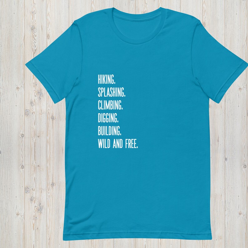 K&ouml;nnte beinhalten: Ein t&uuml;rkisfarbenes T-Shirt mit wei&szlig;em Text, der "HIKING. SPLASHING. CLIMBING. DIGGING. BUILDING. WILD AND FREE." lautet.