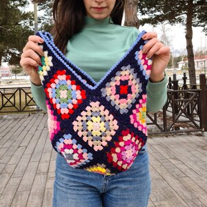 May include: A woman is holding a crocheted tote bag with a colorful granny square pattern. The bag is made with blue, pink, red, yellow, green, and white yarn.