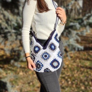 May include: A white crocheted shoulder bag with blue and gray geometric patterns. The bag has a long strap and is worn by a person wearing a white sweater and jeans.