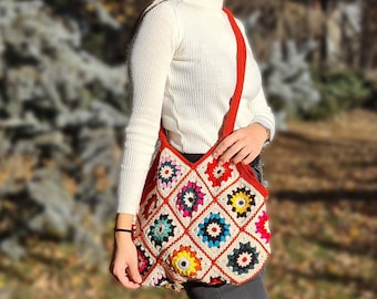 May include: A colorful crochet shoulder bag with a red strap. The bag features a patchwork design with squares of various colors, including red, yellow, blue, and white. The bag is worn over the shoulder.