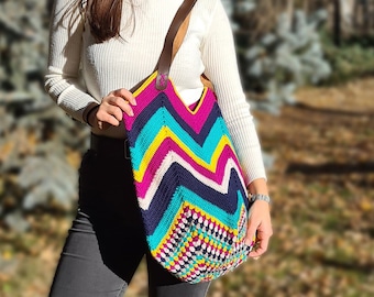 Könnte beinhalten: Eine bunte gehäkelte Tragetasche mit einem Zickzackmuster in den Farben Pink, Blau, Gelb und Weiß. Die Tasche hat einen braunen Lederriemen.