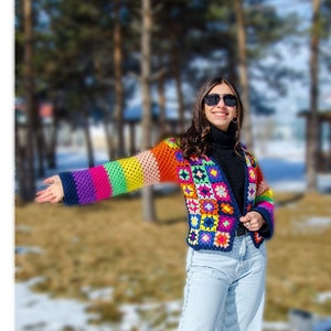 Puede incluir: Una mujer lleva una chaqueta de ganchillo de colores con un patr&oacute;n geom&eacute;trico. La chaqueta tiene rayas de arco&iacute;ris en las mangas y una base negra. Lleva pantalones vaqueros azules y gafas de sol.
