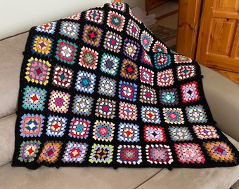 Op de afbeelding: Een zwart gehaakt deken met kleurrijke vierkanten. Het deken is gemaakt van garen en heeft een granny square patroon.