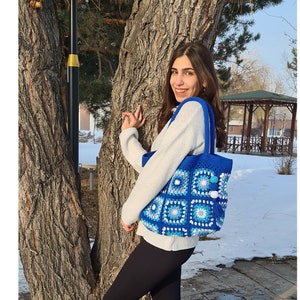 May include: A woman is standing next to a tree wearing black trousers and boots. She is carrying a blue and white crocheted tote bag with pom poms.