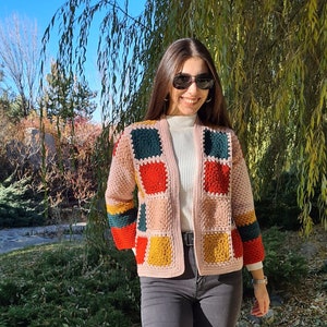May include: A woman wearing a colorful crocheted cardigan with a white turtleneck sweater and dark wash jeans. She is smiling and wearing sunglasses.