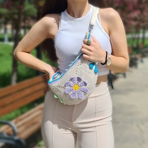 May include: A crocheted flower-shaped fanny pack with a light blue, yellow, and green flower design. The bag is worn over the shoulder of a person wearing a white tank top and beige pants.