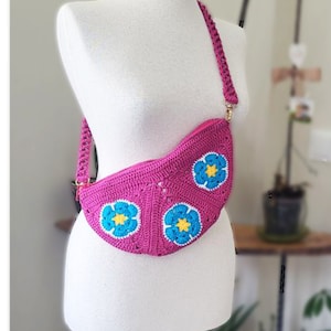 May include: A pink crocheted crossbody bag with a floral design. The bag has three blue flowers with yellow centers.