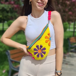 May include: A yellow crocheted fanny pack with colorful flowers and a pink strap. The fanny pack is worn by a person with a white tank top and beige pants.