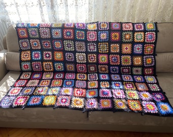Op de afbeelding: Een kleurrijk gehaakt deken met een zwarte achtergrond en vierkanten in verschillende kleuren, waaronder roze, paars, blauw, groen, geel en oranje. Het deken ligt op een bank.