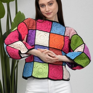 Pode incluir: Um top de crochê colorido com um design de patchwork que apresenta quadrados em vários tons de rosa, verde, azul, laranja e roxo. O top tem um caimento solto e um decote redondo.