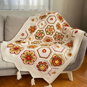 Może przedstawiać: Biały pled szydełkowy z geometrycznym wzorem sześciokątów ozdobionych kolorowymi kwiatami w odcieniach czerwieni, pomarańczy, żółci i zieleni. Pled jest położony na jasnoszarym kanapie z frędzlami zwisającymi z krawędzi.