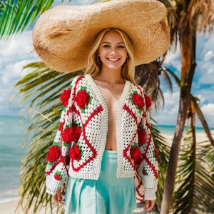 May include: A woman wearing a white crochet cardigan with red roses, a light blue skirt, and a large straw hat. She is standing on a beach with palm trees in the background.