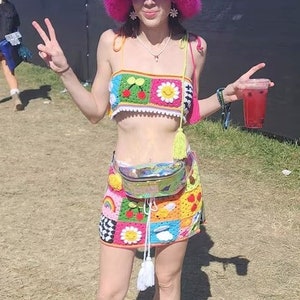 May include: A woman wearing a colourful crochet top and skirt with a pink fluffy hat. She is holding a red drink and smiling.