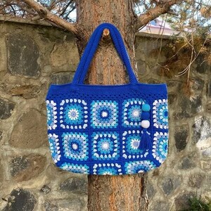 May include: A blue and white crocheted tote bag with a white pom-pom and tassel hanging from the handle. The bag is hanging on a tree trunk.