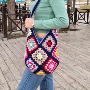 May include: A woman wearing a light green turtleneck sweater and blue jeans is carrying a colorful crocheted tote bag with a blue strap.