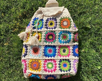 Può includere: Uno zaino all'uncinetto con un motivo geometrico colorato. Lo zaino è realizzato in filato bianco e presenta una varietà di colori, tra cui rosso, arancione, giallo, verde, blu e viola.