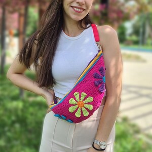 May include: A woman wearing a white tank top and beige pants is standing in a park. She is wearing a pink, yellow, green, and blue crocheted fanny pack with a flower design.