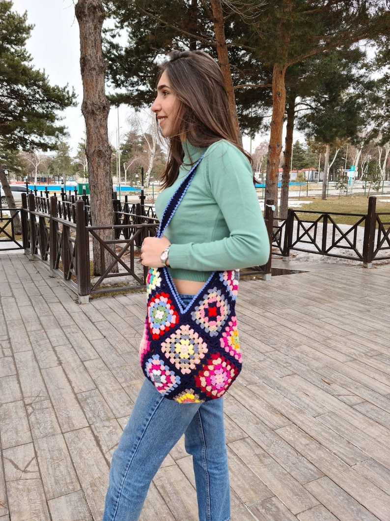 May include: A woman wearing a light green turtleneck sweater and blue jeans is carrying a colorful crocheted tote bag with a blue strap.