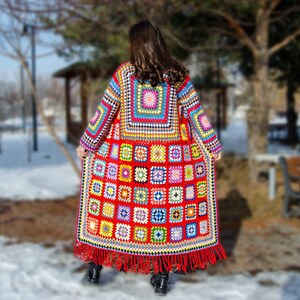 May include: A long, colorful crochet coat featuring a patchwork design with red, yellow, blue, and green squares. The coat has long sleeves and a fringed hem. The person is standing in a snowy park.