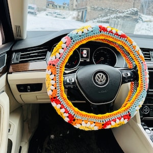 May include: A colorful crocheted steering wheel cover with a multi-colored design of red, orange, yellow, white, and blue. The cover is made of yarn and has a textured surface.