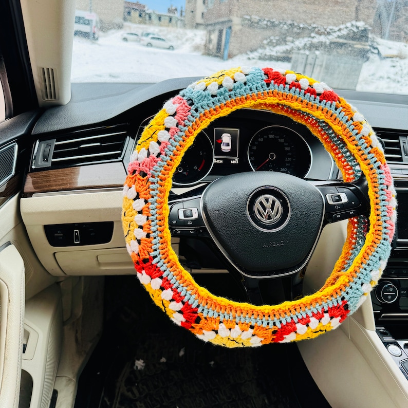 May include: A colorful crocheted steering wheel cover with a multi-colored design of red, orange, yellow, white, and blue. The cover is made of yarn and has a textured surface.